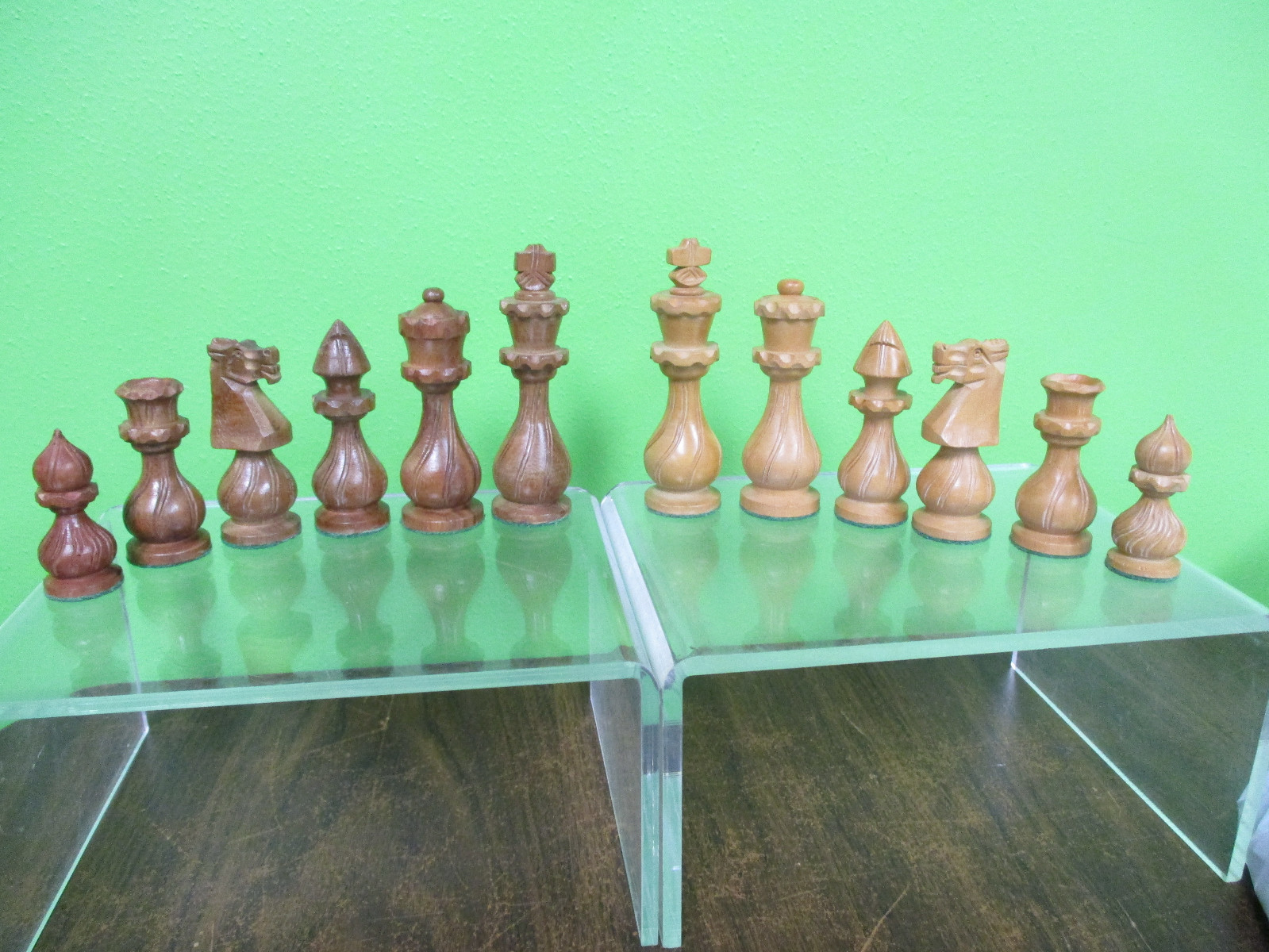 Due serie di pezzi degli scacchi in legno scolpito, marrone scuro e beige chiaro, disposte su basi di vetro trasparente contro una parete verde
